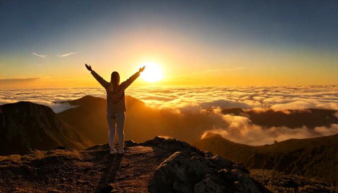 A person standing at the edge of a mountain during sunrise, arms lifted in surrender, with golden light breaking through the clouds